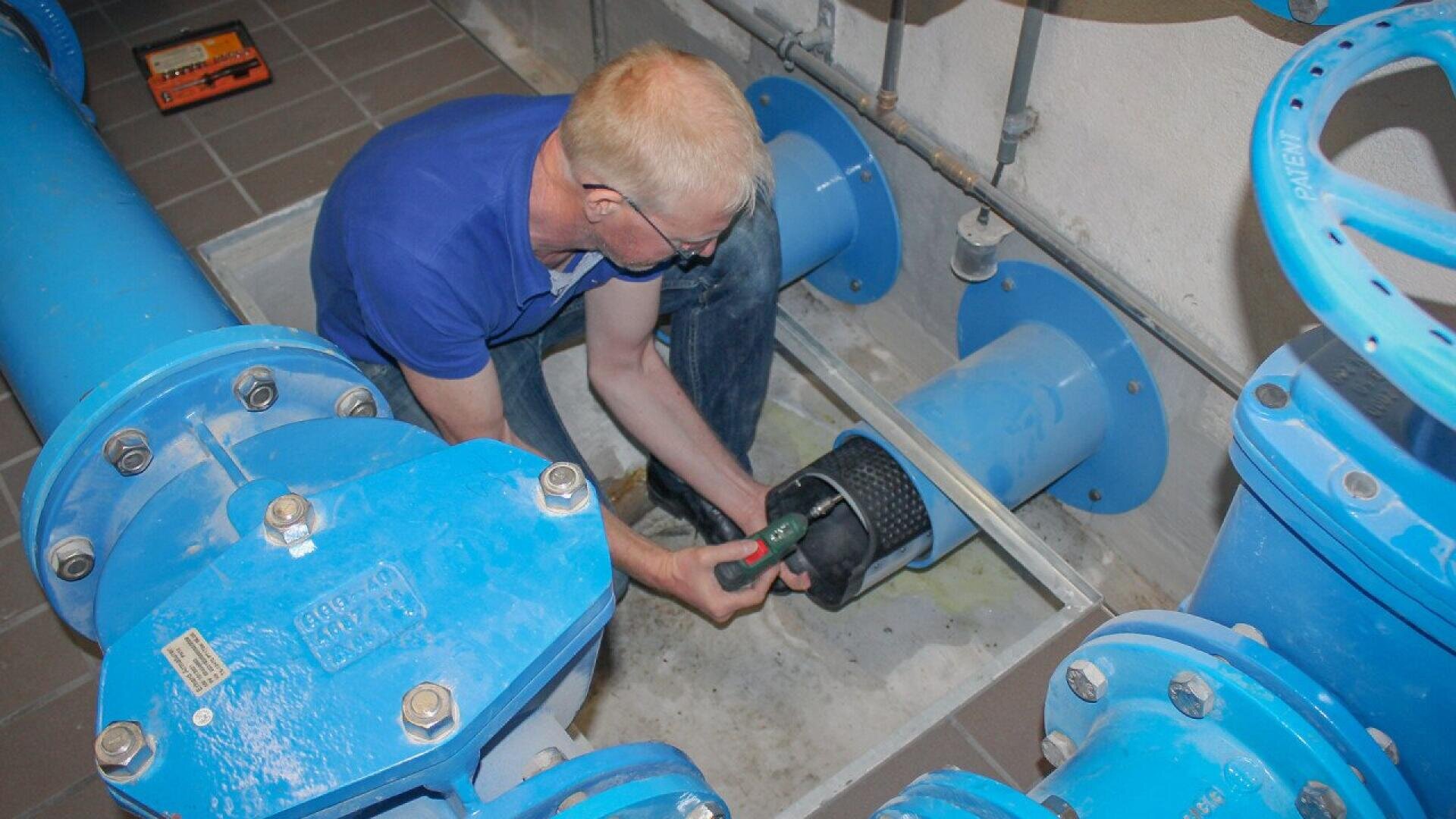 Une personne vêtue d'une chemise bleue et d'un jean est accroupie dans une zone de maintenance, utilisant un outil électrique pour travailler sur un gros tuyau bleu entouré d'autres tuyaux et vannes.