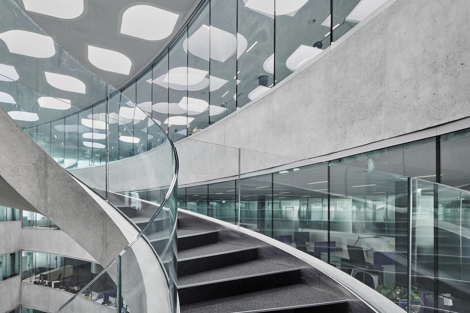 Eine moderne Büroeinrichtung mit einer breiten, geschwungenen Treppe mit Glasgeländer und Betonwänden. Die Decke ist mit geometrischen Lichtpaneelen versehen, und auf mehreren Etagen sind Büros mit Glaswänden zu sehen.
