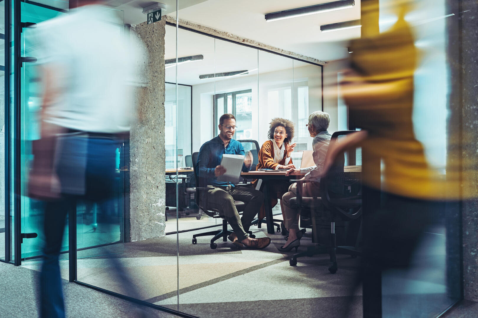 Drei Personen sitzen an einem Tisch in einem modernen Büro mit Glaswänden und diskutieren mit Papieren und Laptops. Draußen laufen verschwommene Gestalten vorbei und schaffen eine geschäftige, dynamische Atmosphäre.