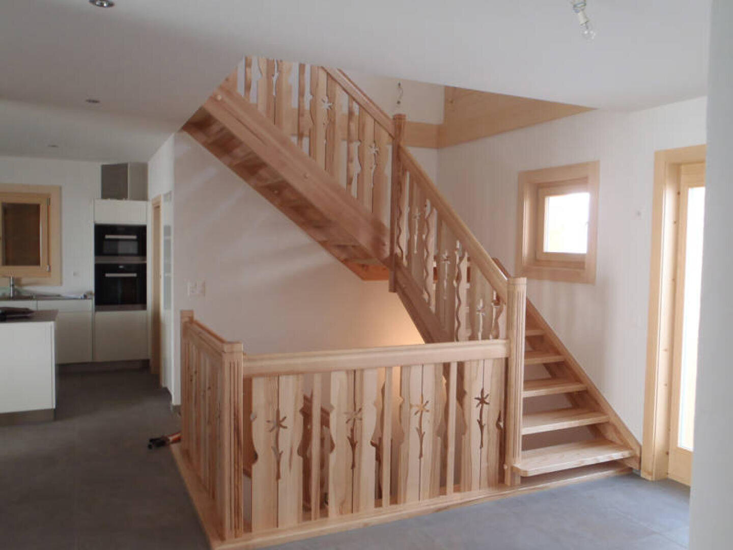 Eine Holztreppe mit dekorativen Geländern und Balustern in einem modernen, hellen Innenraum. Die Treppe führt in ein Obergeschoss und ist von weißen Wänden, hellen Holzverkleidungen und einem gefliesten Boden umgeben.