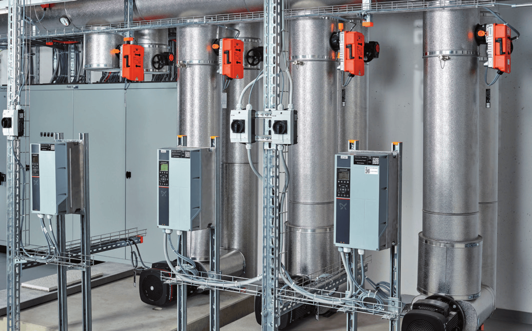 De gros tuyaux métalliques isolés et des servomoteurs orange sont reliés à des tableaux électriques et à des appareils électriques dans une salle des machines industrielle. Le dispositif comprend des câbles, des moteurs et des composants de ventilation ou de chauffage, ventilation et climatisation.