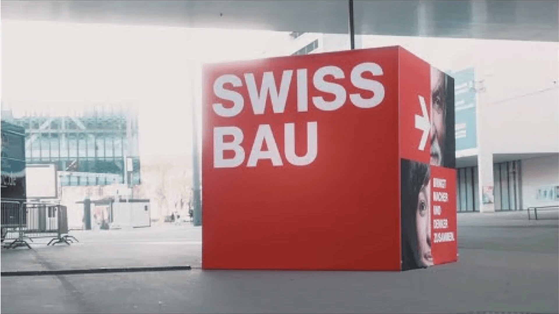 Ein großer roter Würfel mit dem Schriftzug SWISS BAU in fetten weißen Buchstaben ist im Freien in einer modernen städtischen Umgebung aufgestellt. Der Würfel hat Flächen mit Teilbildern von Menschengesichtern und zusätzlichem Text.