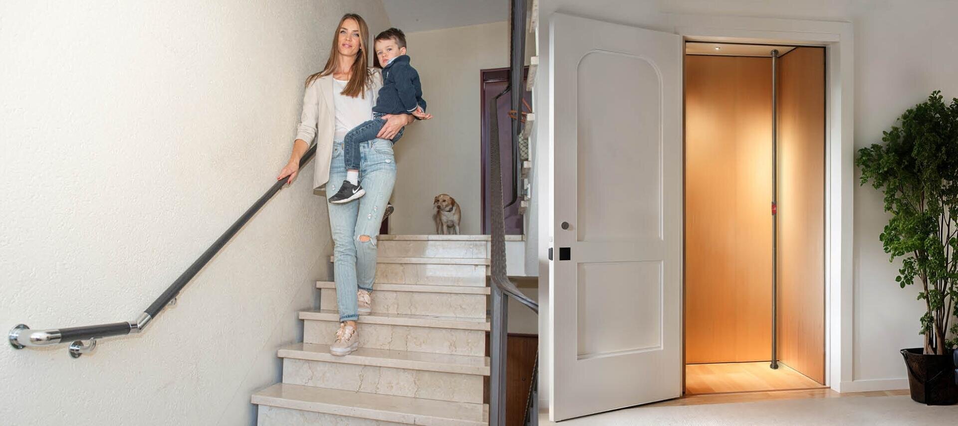 Eine Frau trägt ein kleines Kind eine Treppe mit Handlauf hinunter, während oben ein kleiner Hund wartet; daneben ist ein offener Hausaufzug mit holzgetäfelten Wänden zu sehen.