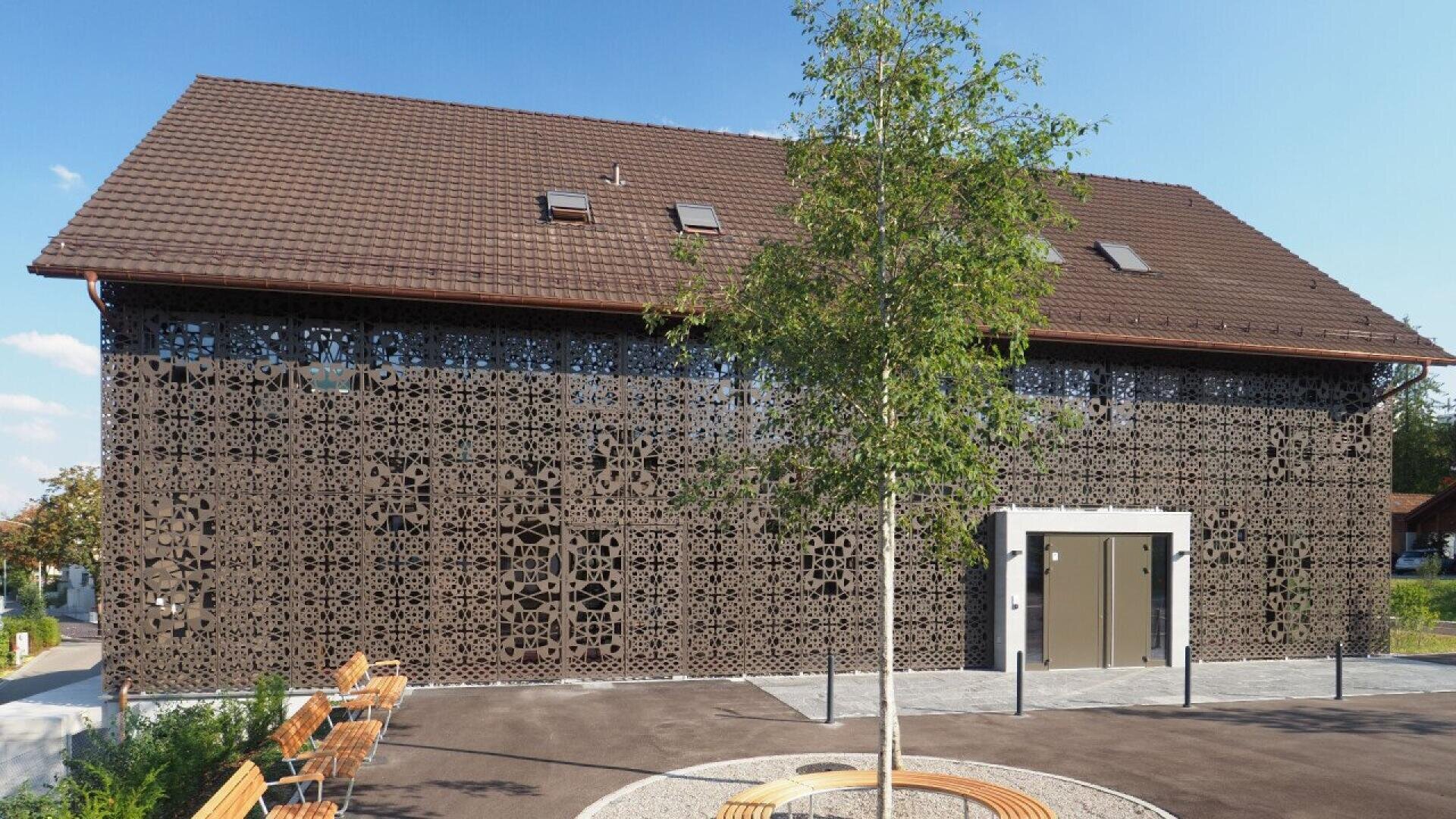 Un bâtiment moderne avec un toit en tuiles brunes et une façade métallique décorative aux motifs géométriques complexes. Un jeune arbre et des bancs se trouvent devant, sous un ciel bleu clair.