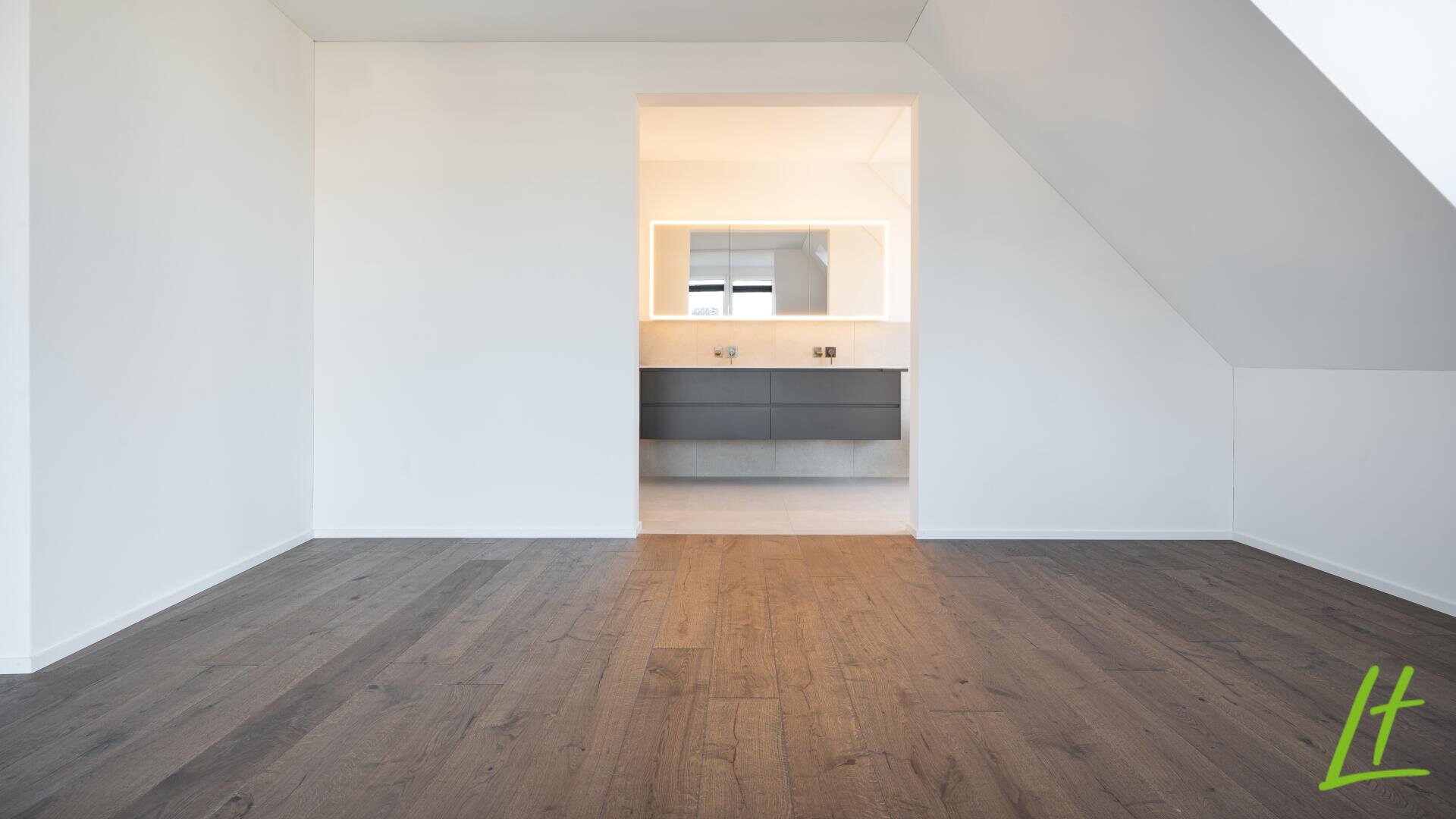 Une pièce moderne et vide avec des murs blancs, un sol en bois foncé et une porte ouverte menant à une salle de bain bien éclairée avec un grand miroir et un meuble-lavabo flottant. Un logo L vert se trouve dans le coin inférieur droit.