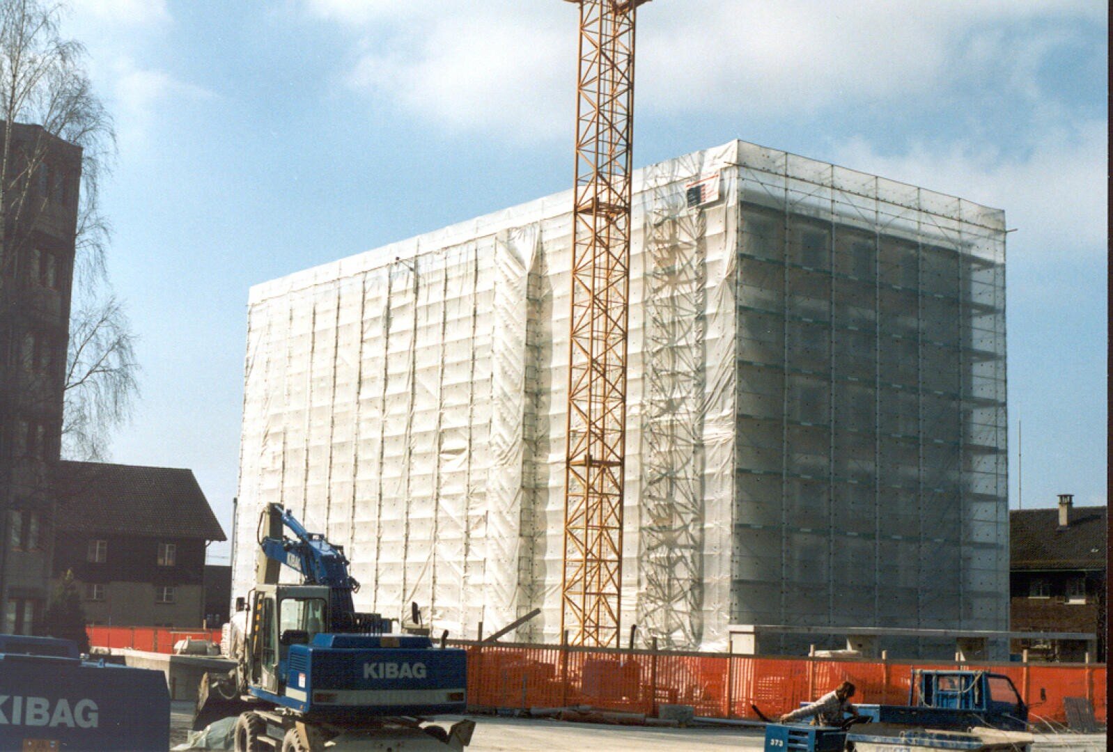 Un grand bâtiment en construction est recouvert d'échafaudages blancs et de filets de protection. Une grande grue jaune se trouve devant, des machines de construction et des matériaux sont posés sur le sol. Le ciel est partiellement nuageux.