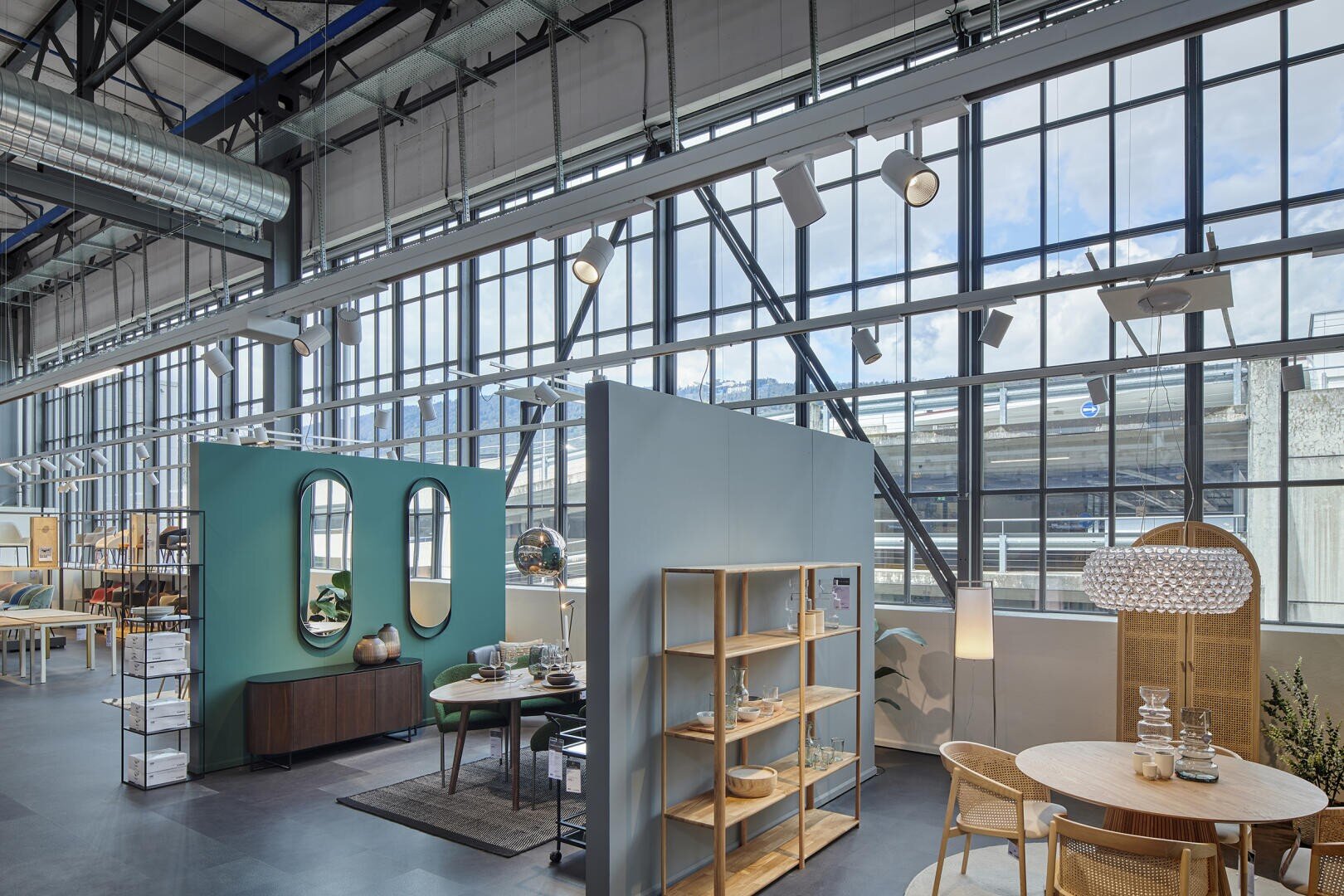 Moderner Ausstellungsraum mit großen Industriefenstern, stilvollen Möbeln wie Tischen, Stühlen, Regalen, dekorativen Spiegeln und Pflanzen, alles in einem hellen, offenen Raum mit hohen Decken angeordnet.
