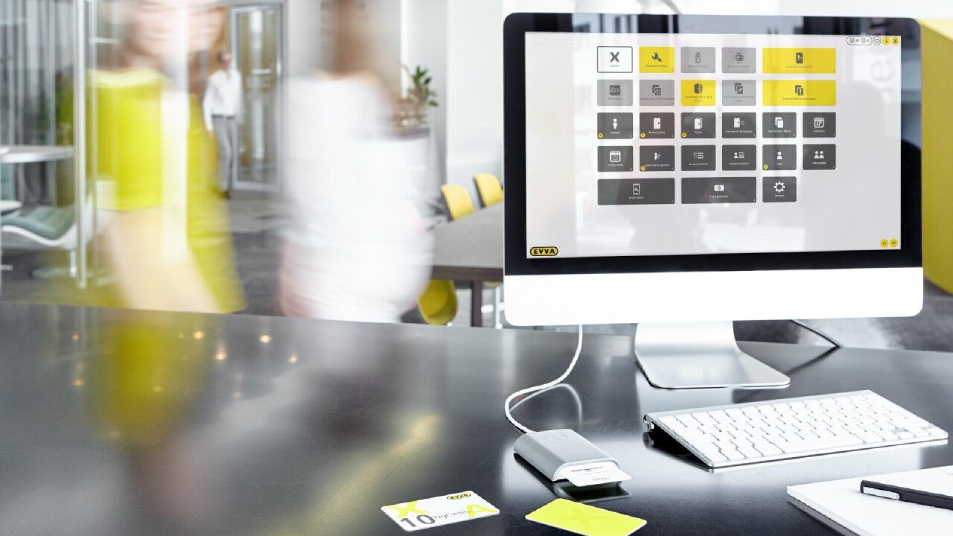 Un bureau moderne avec un ordinateur affichant une interface numérique à l'écran, un clavier, une souris et un lecteur de cartes sur un bureau épuré ; deux personnes floues marchent à l'arrière-plan.