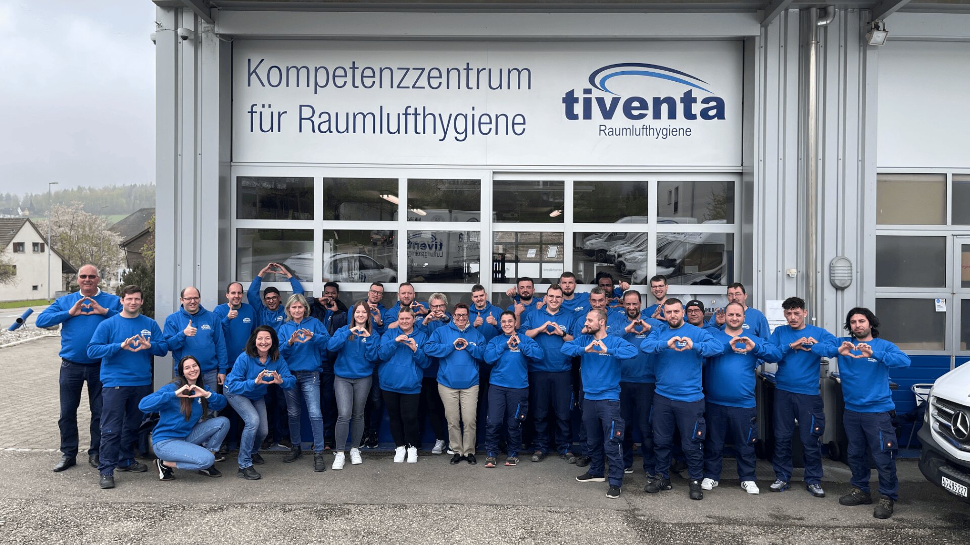 Eine Gruppe von etwa 30 Personen in passenden blauen Pullovern steht vor einem Gebäude mit den Schildern Kompetenzzentrum für Raumlufthygiene und tiventa Raumlufthygiene, posiert und formt mit ihren Händen Herzchen.