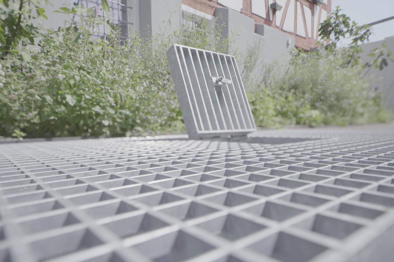 Nahaufnahme einer Metallgitterplattform im Freien, bei der ein quadratisches Gitter als Luke aufgeklappt ist; im Hintergrund sind Grünpflanzen und ein weißes Gebäude zu sehen.