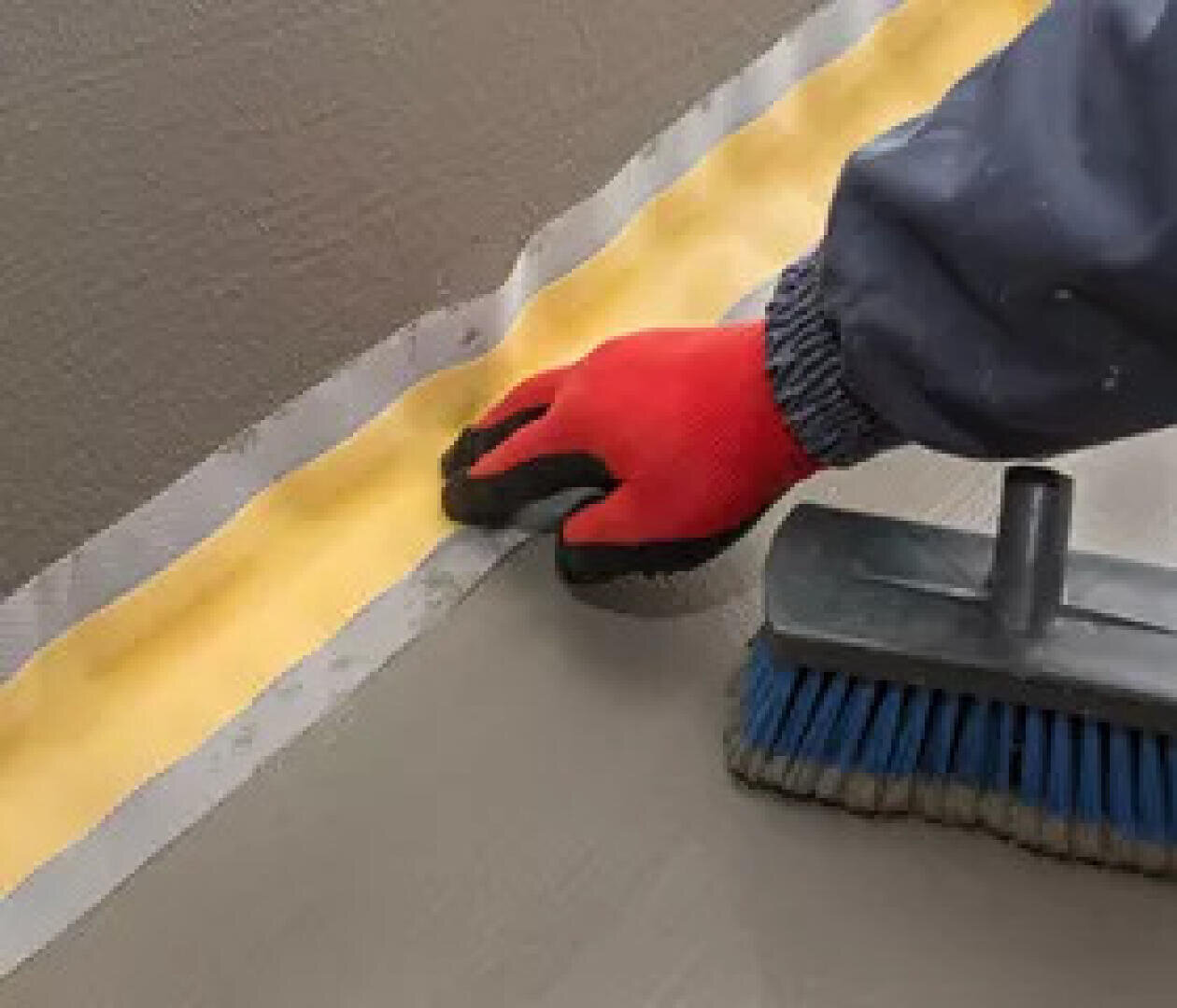 Une personne portant un gant rouge lisse du ruban adhésif jaune sur une surface grise, à côté d'un pinceau à poils bleus, pour préparer la surface à la peinture ou à la vitrification.