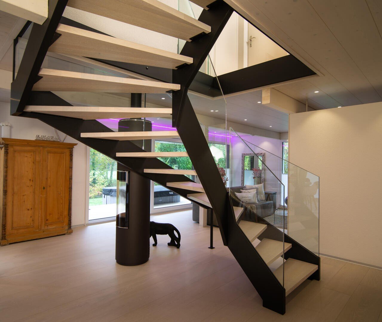 Ein modernes Interieur mit einer schwebenden Treppe mit schwarzen Metallstützen und hellen Holzstufen, Glasgeländern, einem großen Fenster, lila Akzentbeleuchtung, einem Holzschrank und einer schwarzen Hundeskulptur unter der Treppe.