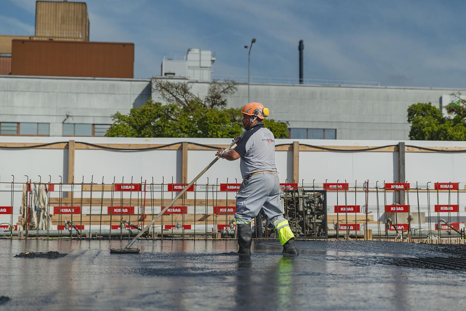 Un ouvrier du bâtiment portant un casque et un équipement de protection lisse du béton mouillé sur un chantier. En arrière-plan, on aperçoit des bâtiments industriels et des barrières de sécurité, le ciel est dégagé.