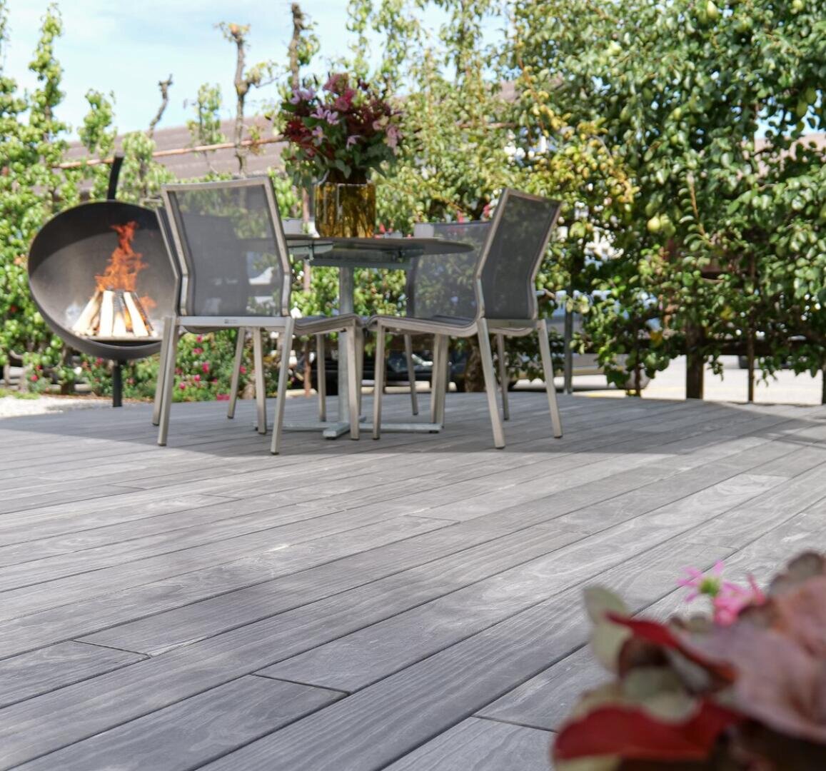 Patio extérieur avec un sol en bois gris, une table ronde avec quatre chaises en treillis, un bouquet de fleurs sur la table, un foyer moderne brûlant à l'arrière-plan, et des plantes entourant la zone.