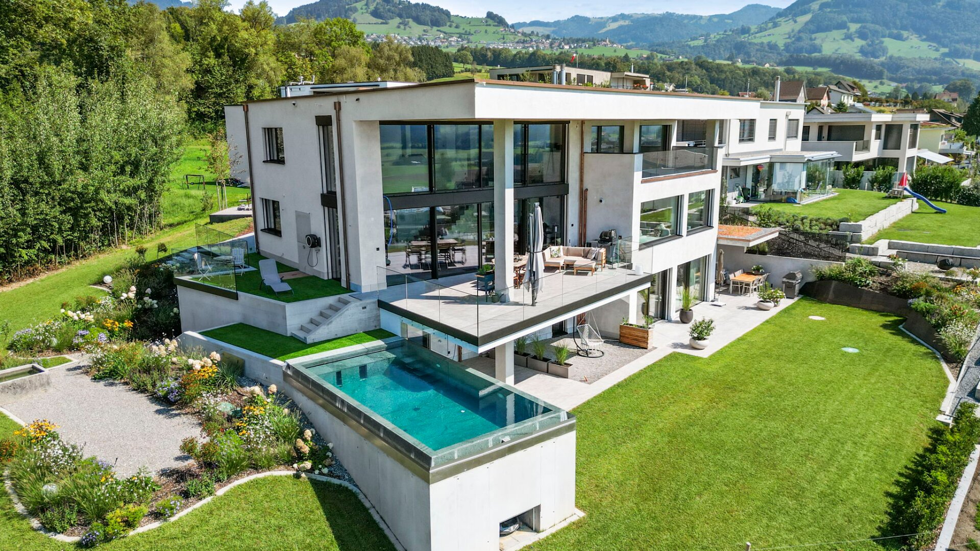 Modernes zweistöckiges Haus mit großen Glasfenstern, mehreren Balkonen und einem Außenpool, umgeben von einem gepflegten Rasen und Garten, mit malerischen grünen Hügeln im Hintergrund.