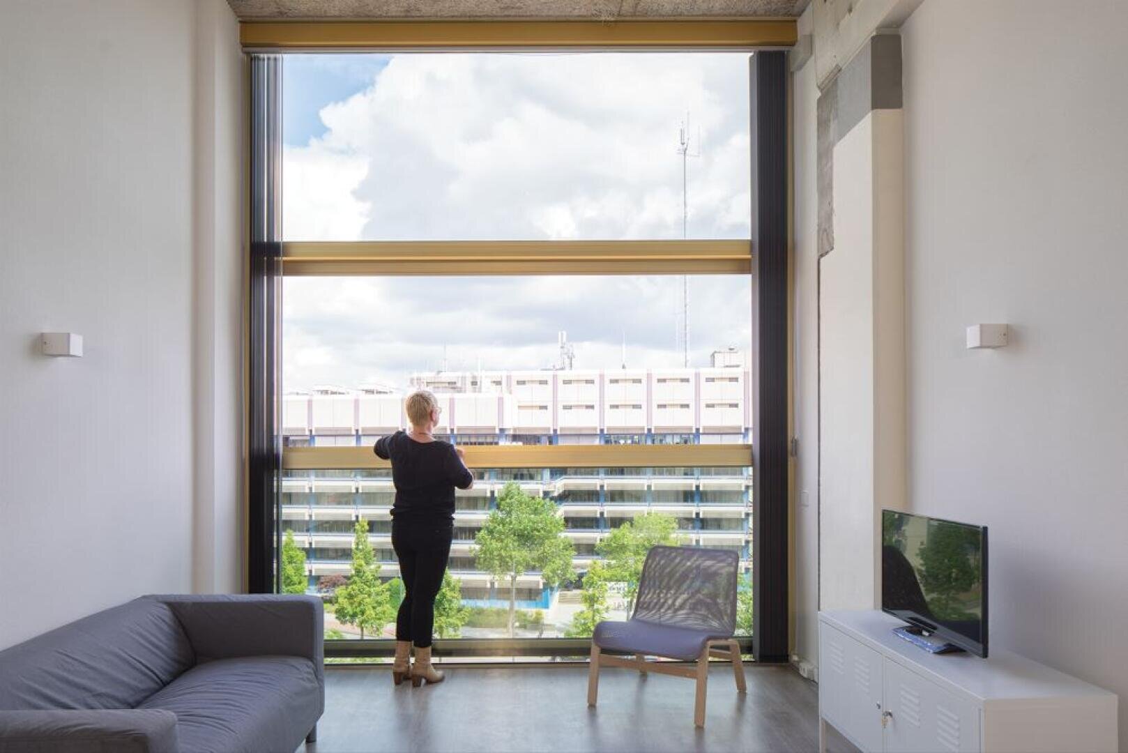 Une personne se tient près d'une grande fenêtre dans un salon moderne et minimaliste, regardant un paysage urbain avec des arbres et un bâtiment sous un ciel partiellement nuageux. Un canapé, une chaise et une télévision se trouvent dans la pièce.