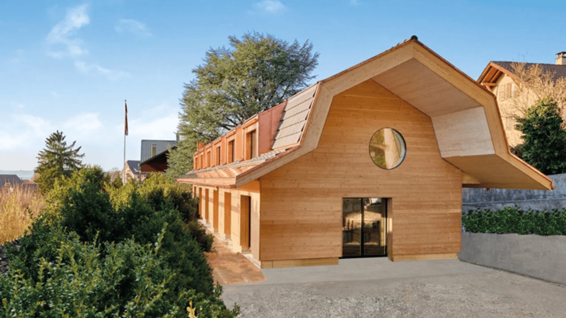 Une maison moderne en bois avec un toit caractéristique de type grange, une fenêtre circulaire au-dessus de portes vitrées, et entourée de verdure par une journée ensoleillée.