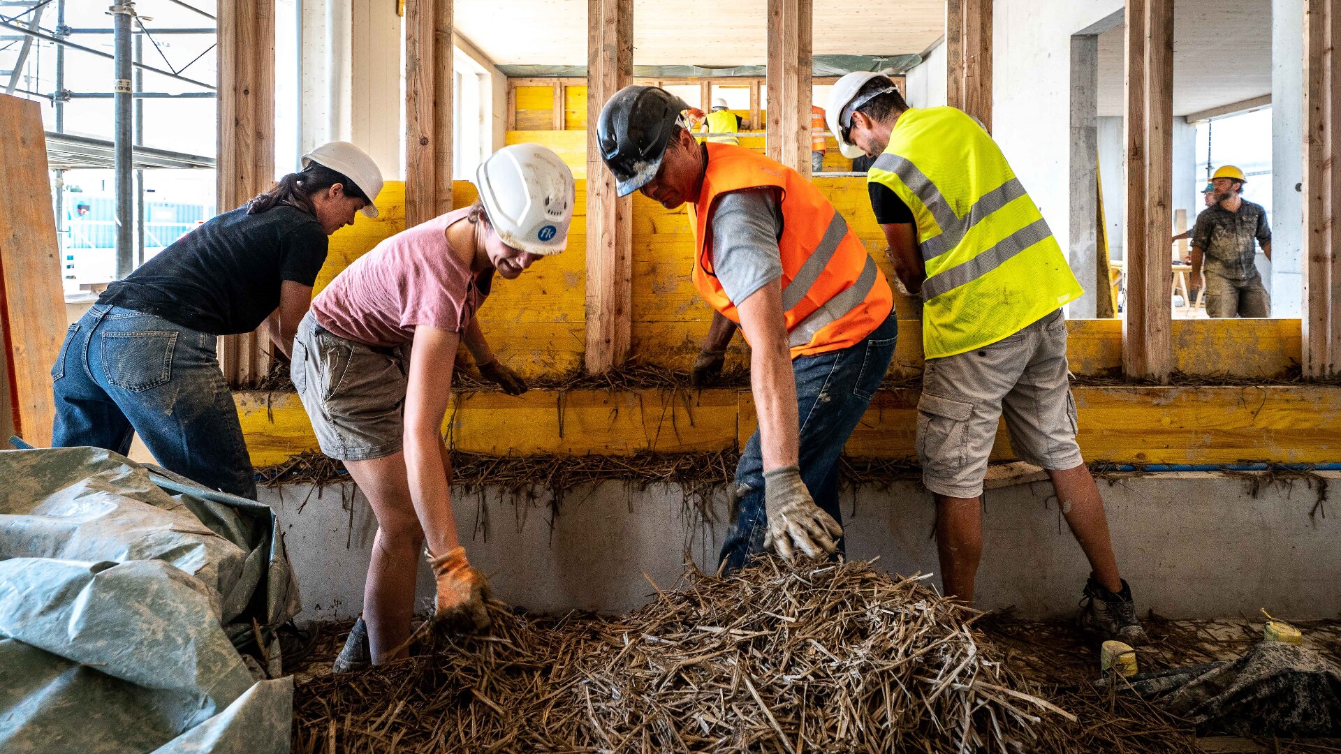 Vier Bauarbeiter mit Schutzhelmen und Sicherheitswesten stecken Stroh in ein Holzgerüst auf einer Baustelle. Das Sonnenlicht beleuchtet die Szene, und um sie herum sind Werkzeuge und Baumaterialien zu sehen.