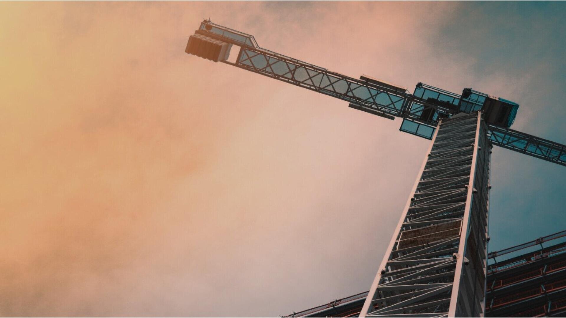 Vue rapprochée d'une grue de construction prise de dessous dans un ciel aux douces nuances d'orange et de bleu.