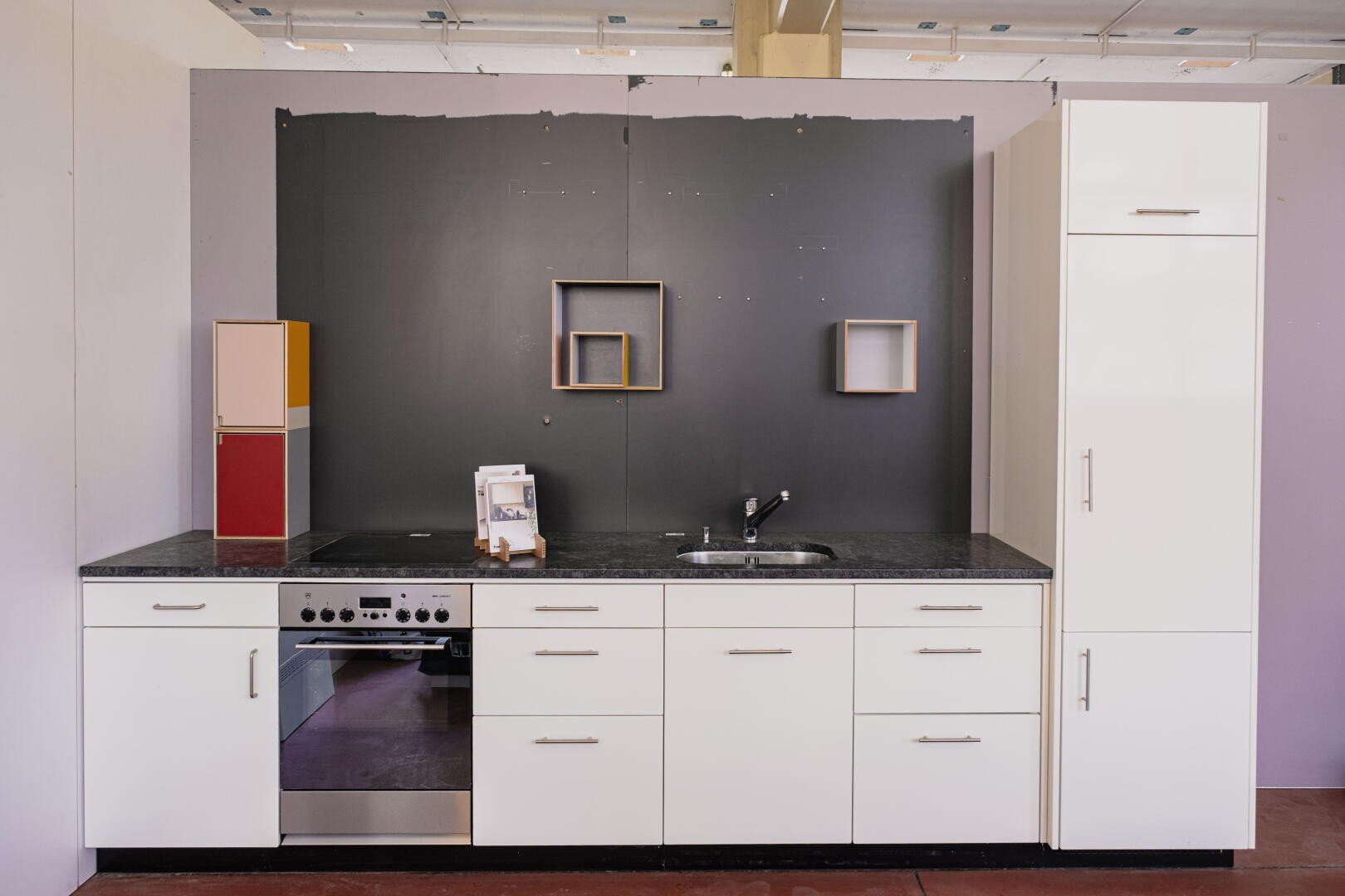 Moderne Küche mit weißen Schränken, schwarzer Arbeitsplatte, eingebautem Ofen, Edelstahlspüle und geometrischen Regalen an einer dunklen Akzentwand. Die Küche wirkt minimalistisch und aufgeräumt.