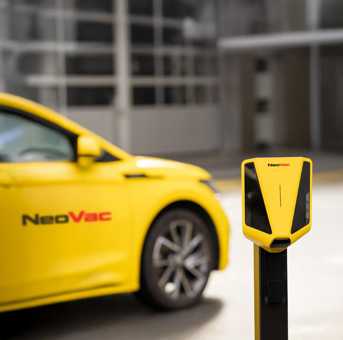 Ein gelbes Elektroauto der Marke NeoVac ist neben einer passenden gelben NeoVac-Ladestation geparkt, mit einem unscharfen Gebäude im Hintergrund.