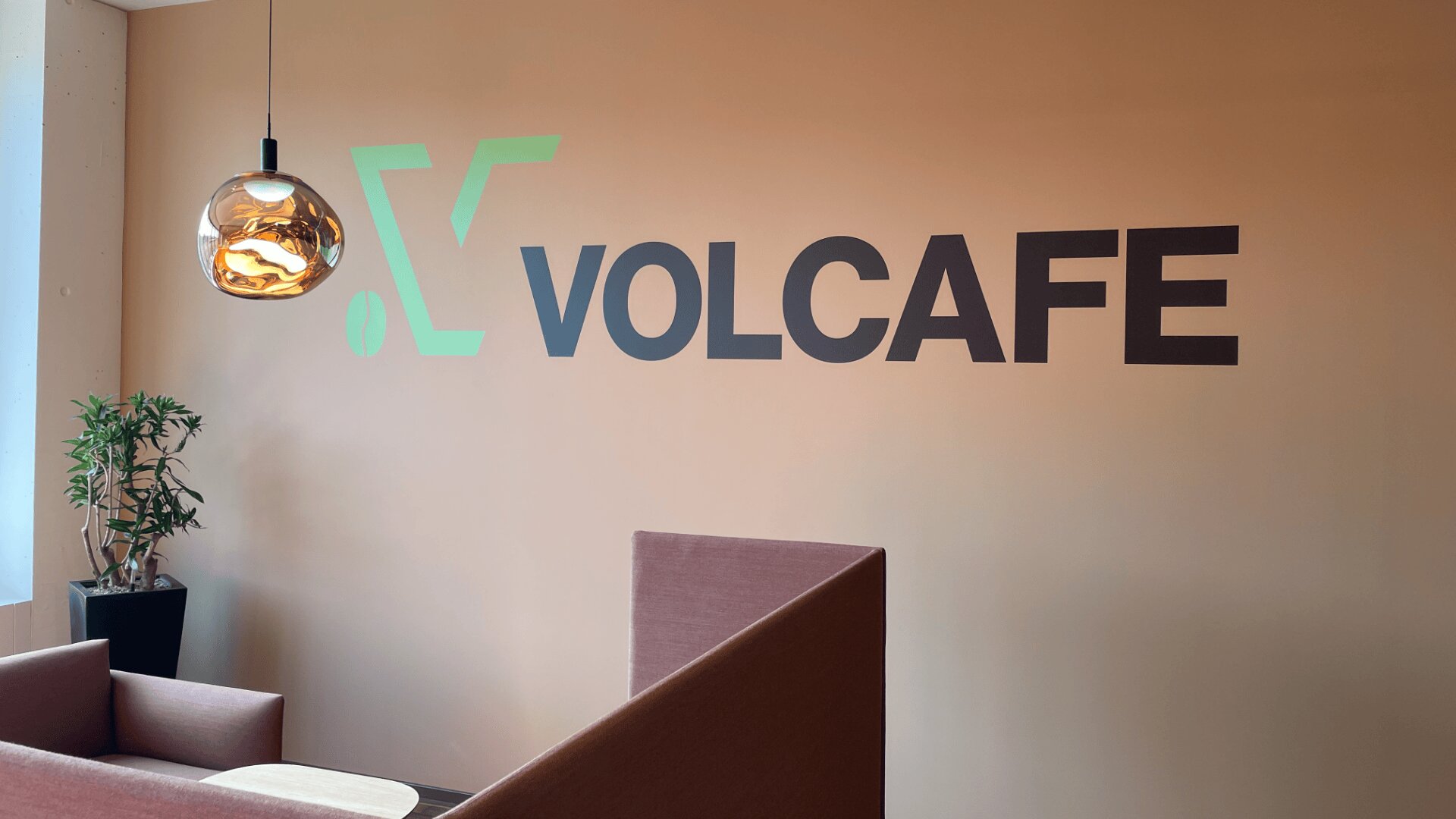Espace de bureau moderne avec un mur couleur pêche affichant le logo VOLCAFE en grandes lettres noires et vertes. Un luminaire suspendu, une plante verte en pot et des sièges bruns sont également visibles.