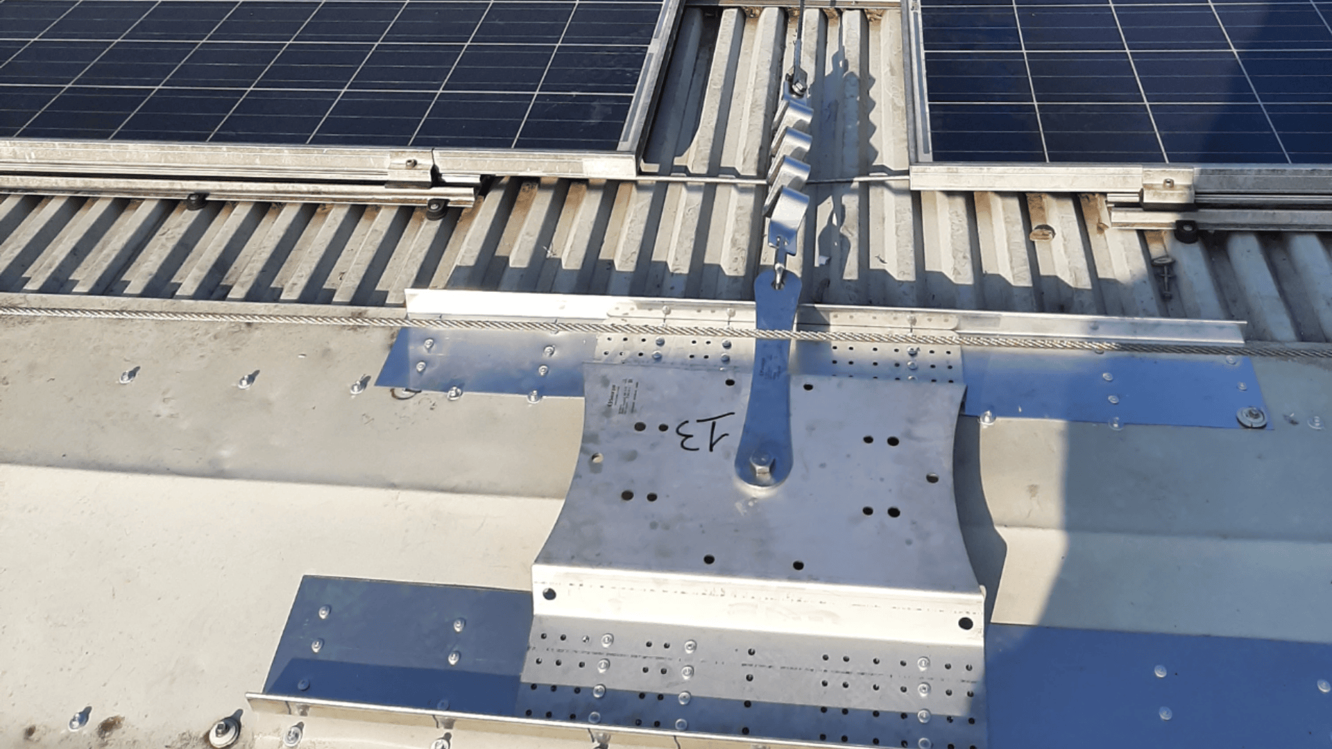 Nahaufnahme einer Metallhalterung, die an einem Wellblechdach befestigt ist, mit einem Teil eines Solarmodulfelds, das am oberen Bildrand zu sehen ist. Das Sonnenlicht wirft Schatten auf die Komponenten.