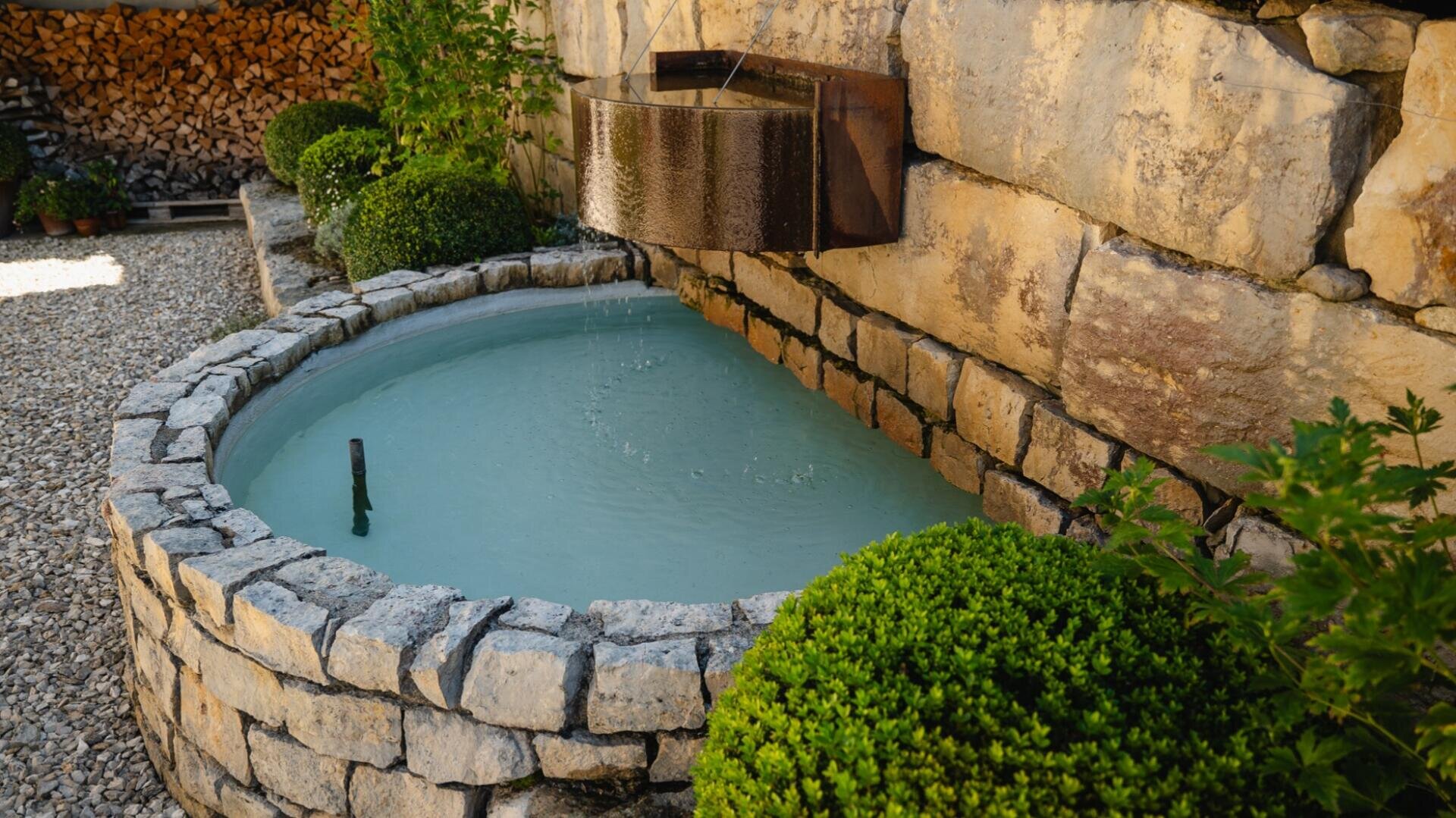 Ein rundes steinernes Wasserbecken mit klarem Wasser steht neben einer Steinmauer in einem Garten, darüber befindet sich ein Metallauslauf. In der Nähe sind grüne Sträucher und ein Stapel Brennholz zu sehen.