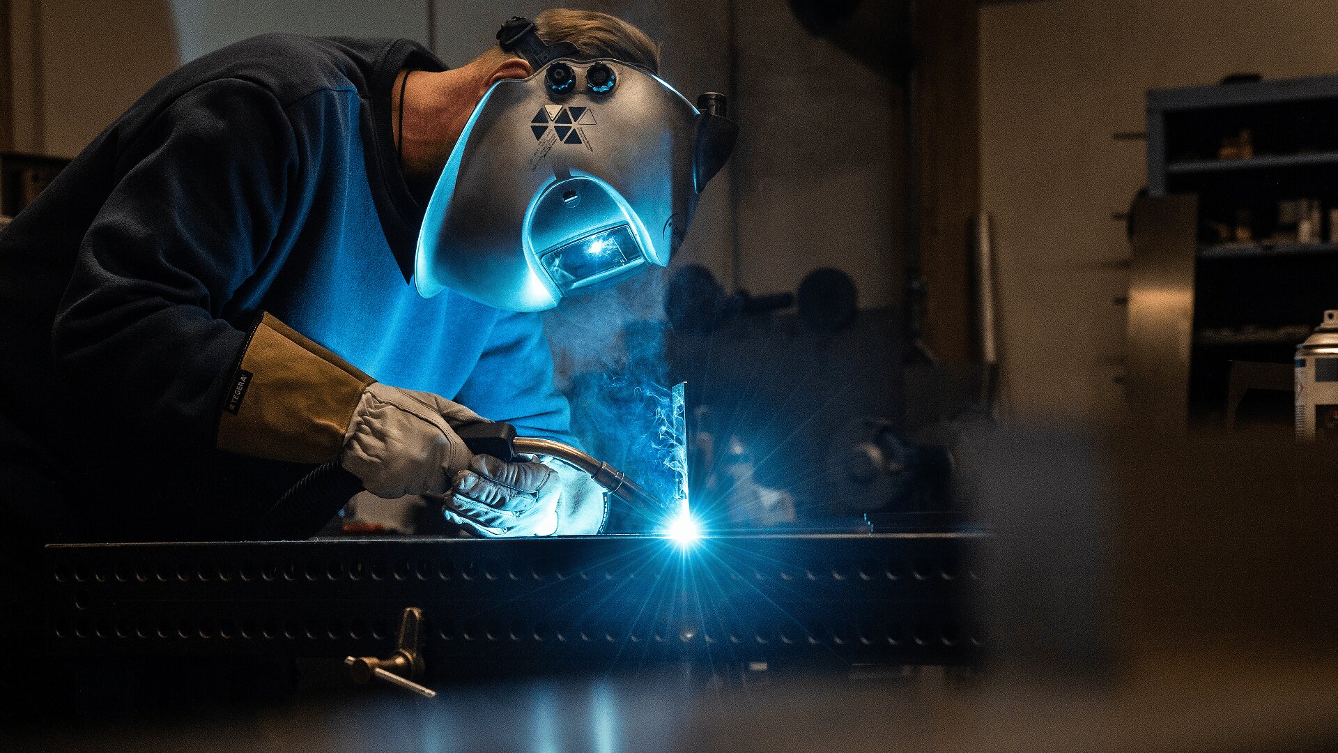 Eine Person, die einen Schweißerhelm und Schutzhandschuhe trägt, schweißt Metall auf einer Werkbank, wobei helle Funken und Rauch vom Schweißvorgang sichtbar sind. Im Hintergrund sind Werkstattausrüstung und Regale zu sehen.