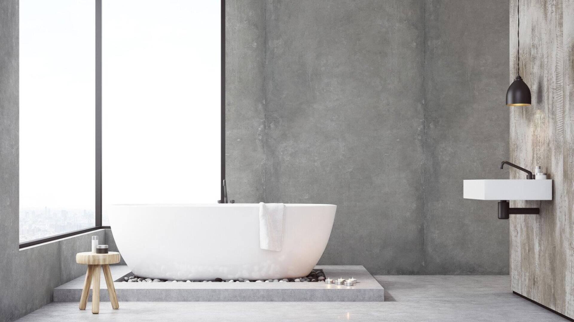 Salle de bains moderne et minimaliste avec une grande baignoire blanche autoportante près d'une fenêtre allant du sol au plafond, un petit tabouret en bois avec des articles de toilette, un lavabo fixé au mur, des murs et un sol en béton.