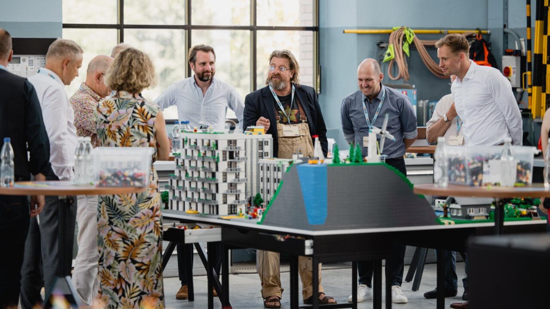 Un groupe de personnes se tient autour d'une table présentant une maquette architecturale détaillée et discute dans une pièce moderne et lumineuse avec de grandes fenêtres et diverses fournitures en arrière-plan.