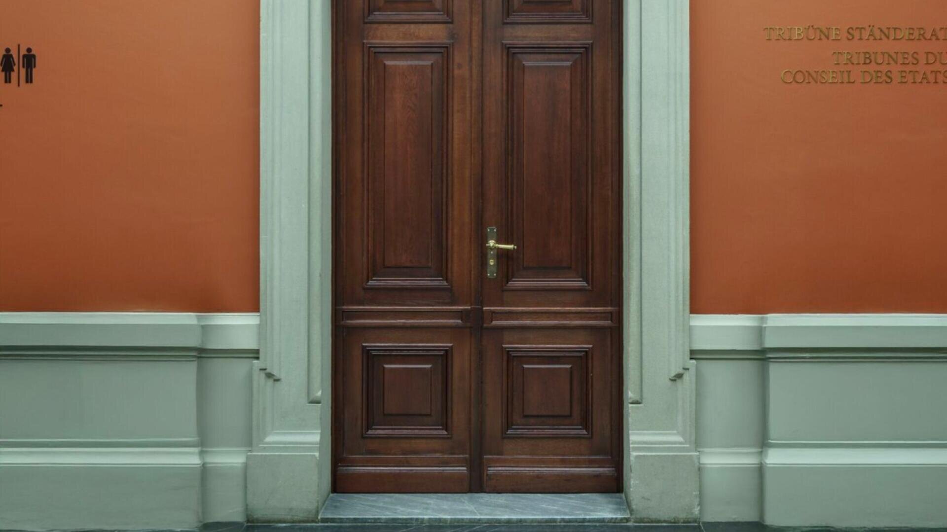 Eine große hölzerne Doppeltür mit Messinggriff in einem Steinrahmen vor einer orangefarbenen Wand mit Schildern und Toilettensymbolen auf beiden Seiten. Die Wand hat einen weißen unteren Teil.