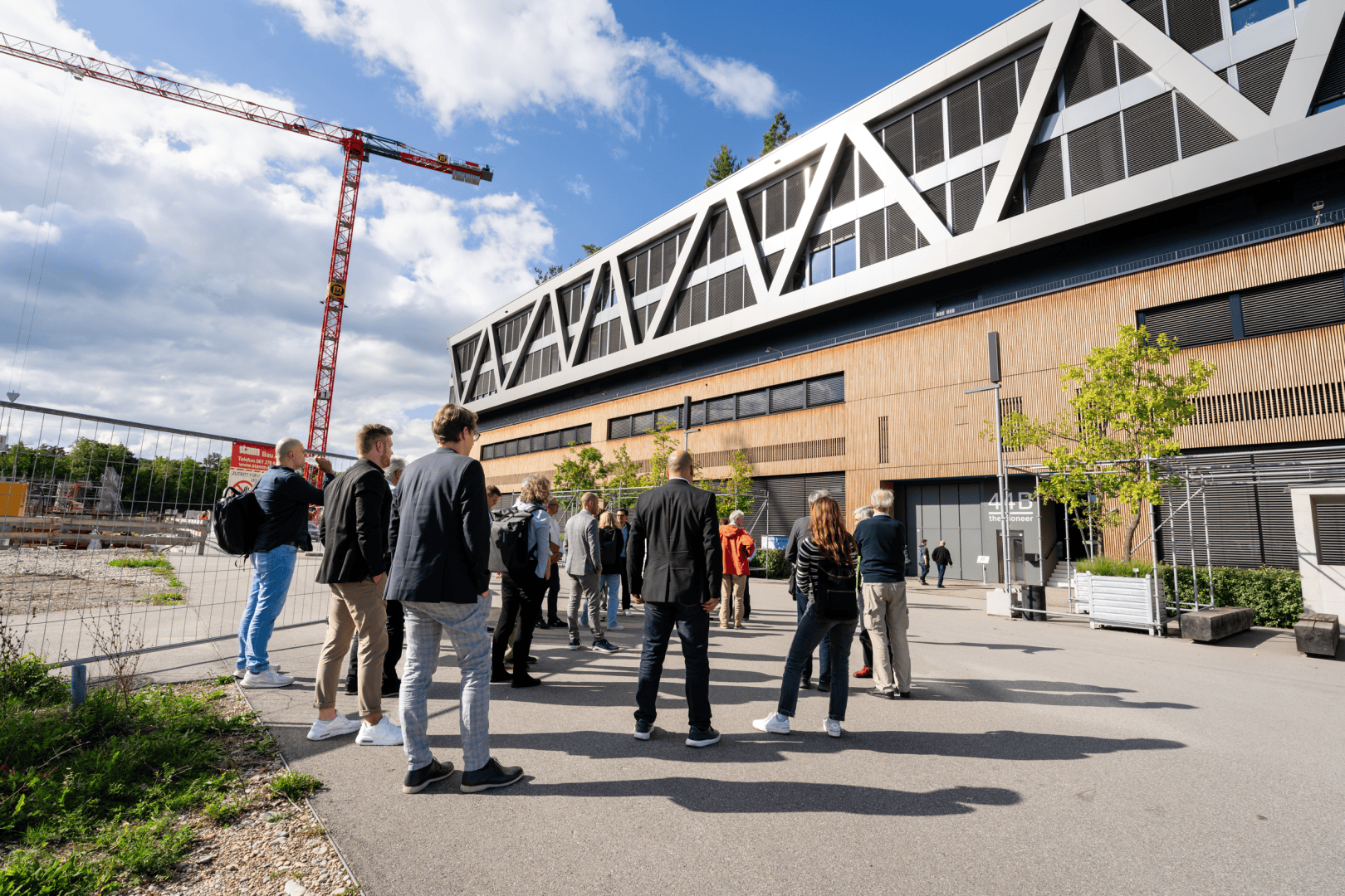 Eine Gruppe von Menschen steht vor einem modernen Gebäude mit geometrischen weißen Akzenten, in der Nähe einer Baustelle mit einem roten Kran unter einem teilweise bewölkten Himmel.