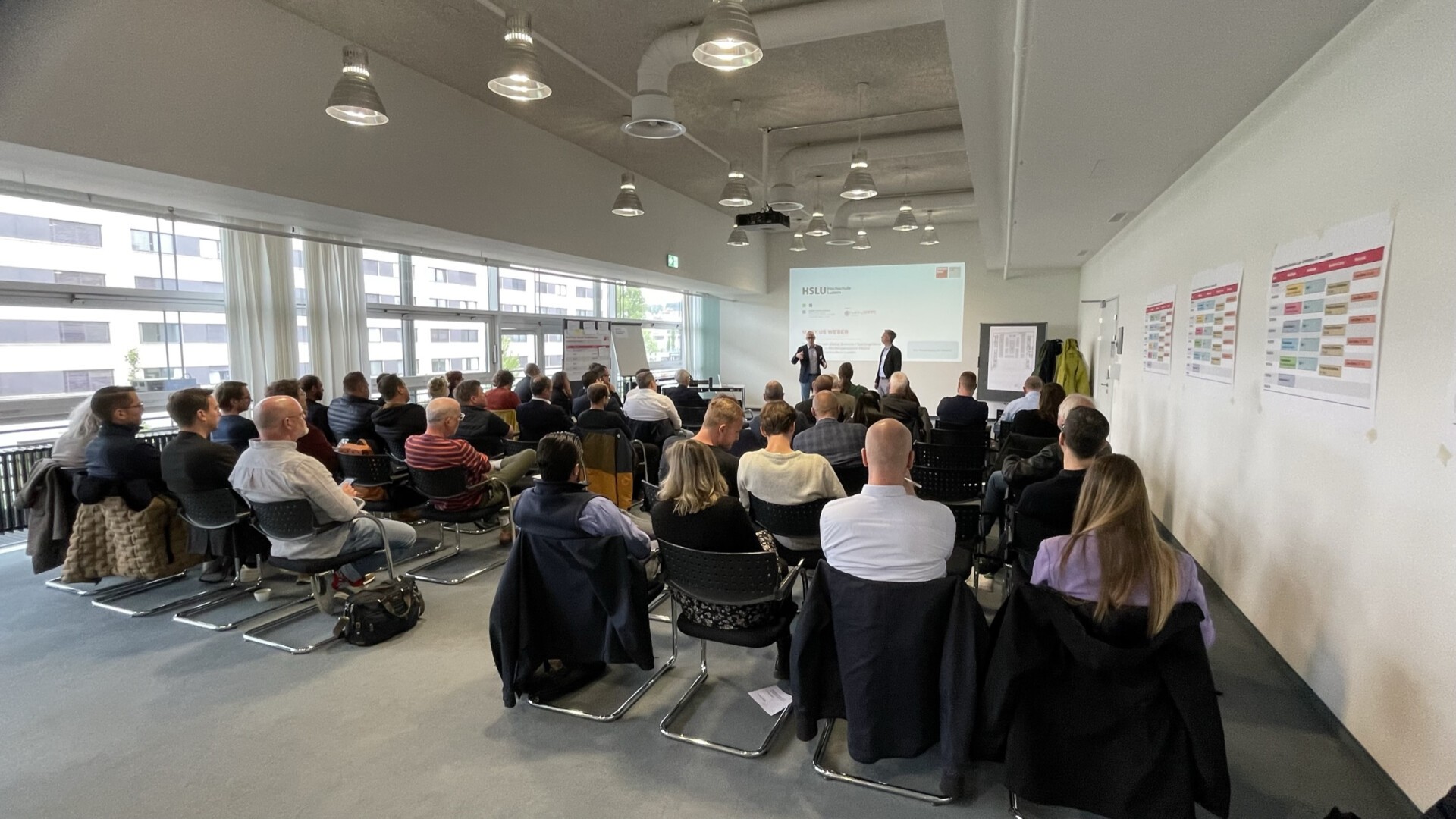 Eine Gruppe von Menschen sitzt in Reihen vor einem Vortragenden im vorderen Teil eines modernen, gut beleuchteten Konferenzraums. Zwei Sprecher stehen neben einer Leinwand, auf der eine Präsentation läuft. An der Wand hängen Diagramme.