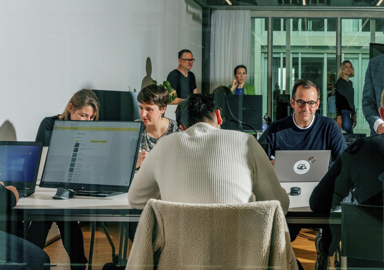 Eine Gruppe von Personen arbeitet an einem großen Tisch mit Laptops in einem modernen Büro, während andere im Hintergrund stehen und sich unterhalten. Die Atmosphäre wirkt kollaborativ und konzentriert.
