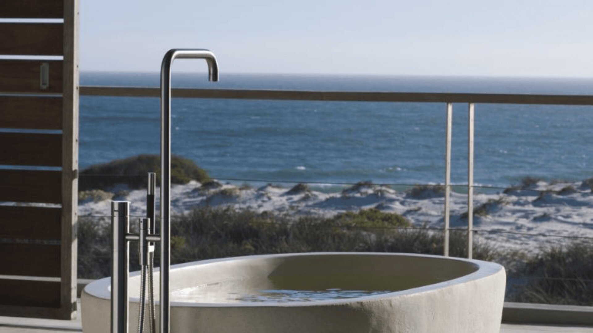 Eine moderne, mit Wasser gefüllte Badewanne steht auf einer Terrasse mit Blick auf die Sanddünen und das Meer, mit einem eleganten Metallhahn und einem klaren blauen Himmel im Hintergrund.