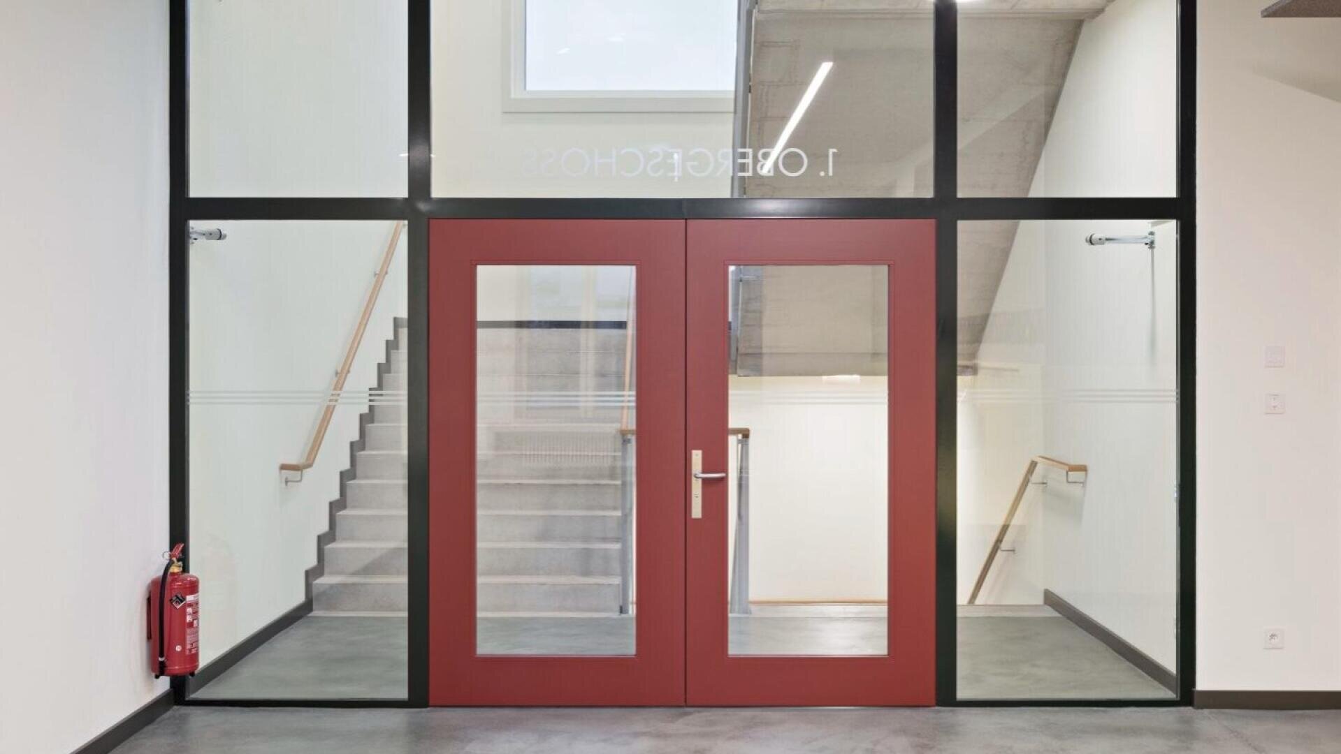 Une cage d'escalier moderne avec une double porte rouge et de grandes vitres qui mènent à un escalier. Un extincteur est placé sur le mur de gauche et une fenêtre au-dessus de la porte laisse entrer la lumière du jour.