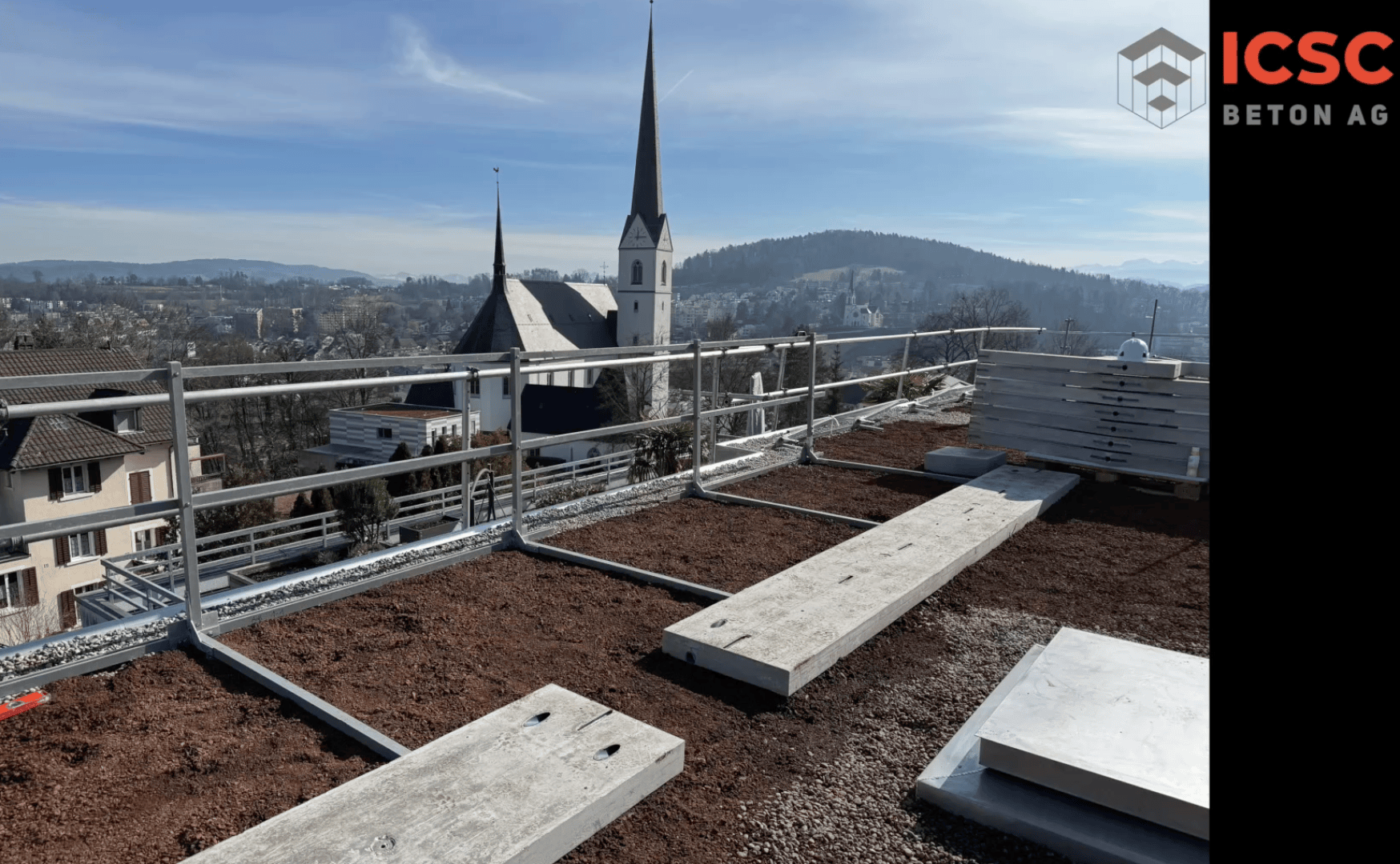 Eine Baustelle auf einem Dach mit Metallgeländern, Betonplatten und Erde. Im Hintergrund sind Häuser, Bäume, Hügel und eine Kirche mit zwei hohen Kirchtürmen zu sehen. In der rechten oberen Ecke ist das Logo der ICSC Beton AG zu sehen.