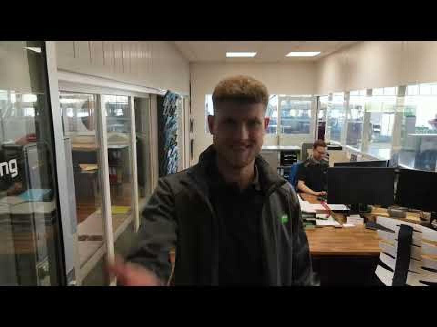 Un homme souriant se tient au premier plan d'un espace de bureau où l'on peut voir des bureaux, des papiers et des ordinateurs. Une autre personne travaille à un bureau à l'arrière-plan, près de grandes fenêtres.