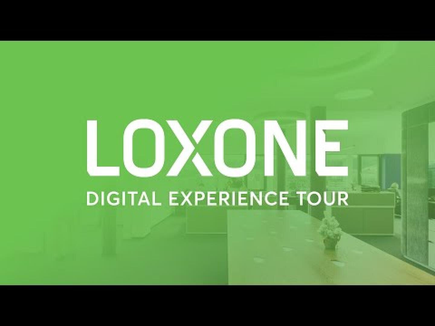 Ein grünes Overlay mit dem weißen Text LOXONE DIGITAL EXPERIENCE TOUR bedeckt einen verschwommenen Hintergrund eines modernen Büro- oder Ausstellungsraums.
