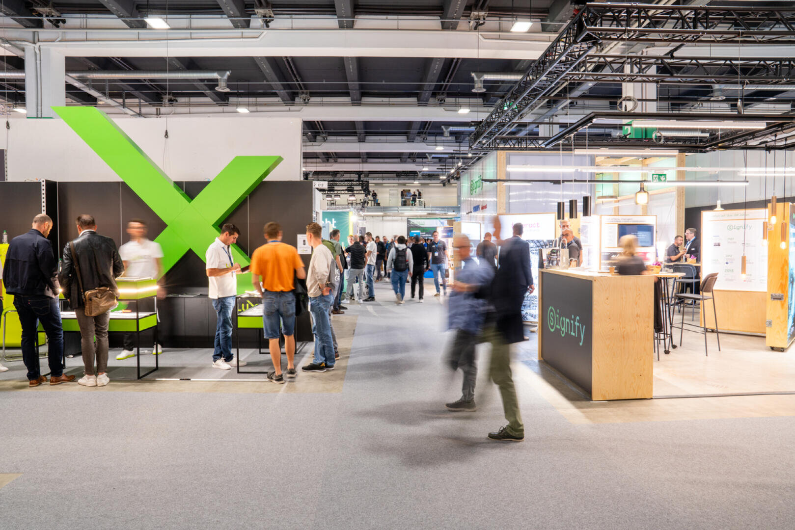 Auf einer hell beleuchteten, modernen Messe oder Ausstellung mit verschiedenen Ständen und Ausstellungsflächen, darunter eine große grüne X-Konstruktion und mehrere Informationsschalter, bewegen sich die Besucher umher und kommen miteinander ins Gespräch.