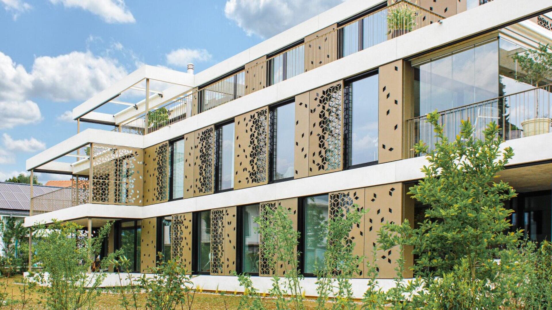 Bâtiment moderne avec de grandes baies vitrées, des panneaux beiges aux découpes irrégulières et des balcons privés. L'environnement comprend des arbustes verts et des petits arbres sous un ciel partiellement nuageux.