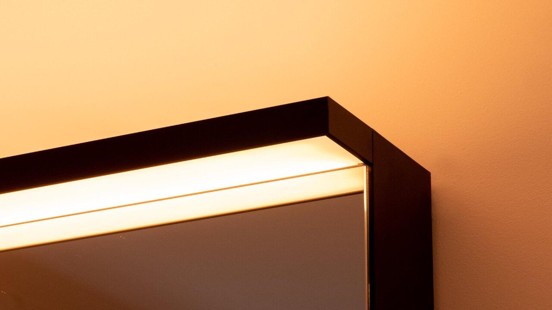 Gros plan sur le coin supérieur d'un miroir rectangulaire au cadre noir, éclairé par une lumière chaude encastrée sur un mur couleur pêche.