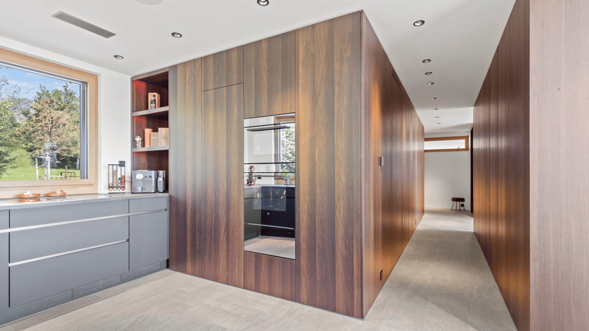 Moderne Küche mit eleganten holzgetäfelten Schränken, eingebautem Backofen, grauen Unterschränken, einem großen Fenster, das natürliches Licht hereinlässt, und einem langen Flur mit hellem Bodenbelag.