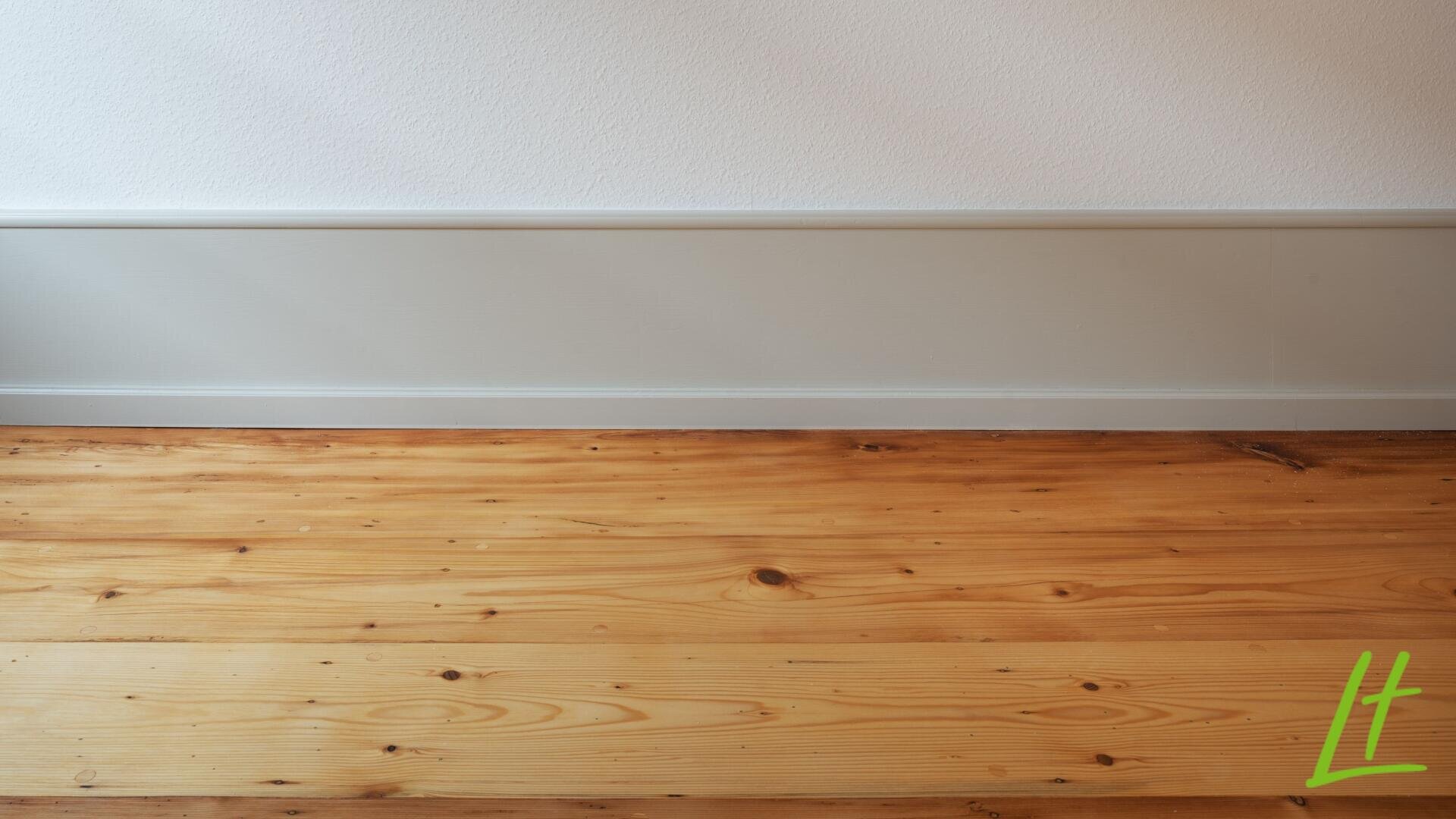 Un plancher en bois clair rencontre un mur blanc avec une plinthe gris clair ; dans le coin inférieur droit se trouve un logo L vert.