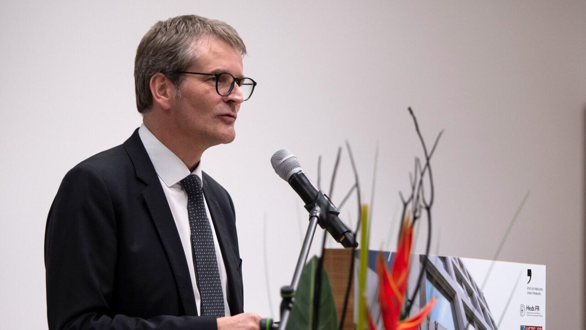 Un homme en costume-cravate parle dans un microphone sur un podium, avec des plantes décoratives et une affiche au premier plan. L'arrière-plan est uni et de couleur claire.