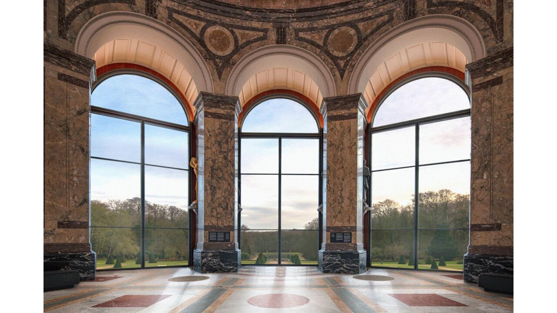 De grandes fenêtres cintrées avec des colonnes en marbre encadrent une vue sur un jardin paysager avec des arbres. L'intérieur élégant présente des murs en marbre ornés et un sol à motifs. La lumière du jour pénètre doucement par les grandes fenêtres.