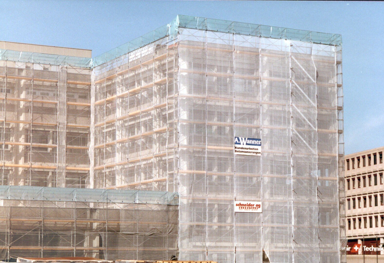 Un grand bâtiment avec des échafaudages et des filets de protection sur lesquels sont fixées des enseignes d'entreprises, le tout sur fond de ciel bleu éclatant.