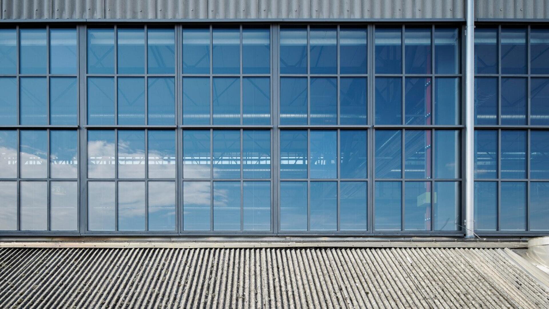De grandes fenêtres de style industriel avec des cadres métalliques noirs reflètent le ciel bleu et les nuages au-dessus d'un toit en tôle ondulée sur un bâtiment moderne.