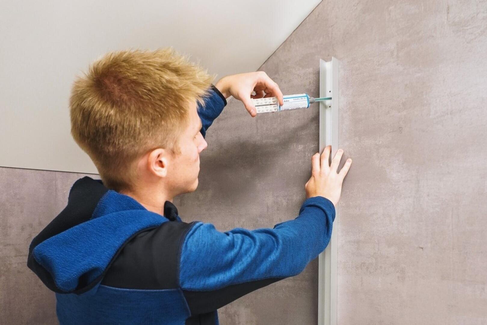 Une personne vêtue d'une veste bleue installe un rail vertical sur un mur, en utilisant un tournevis pour le fixer à l'aide de vis. Le mur est de couleur neutre, beige clair.